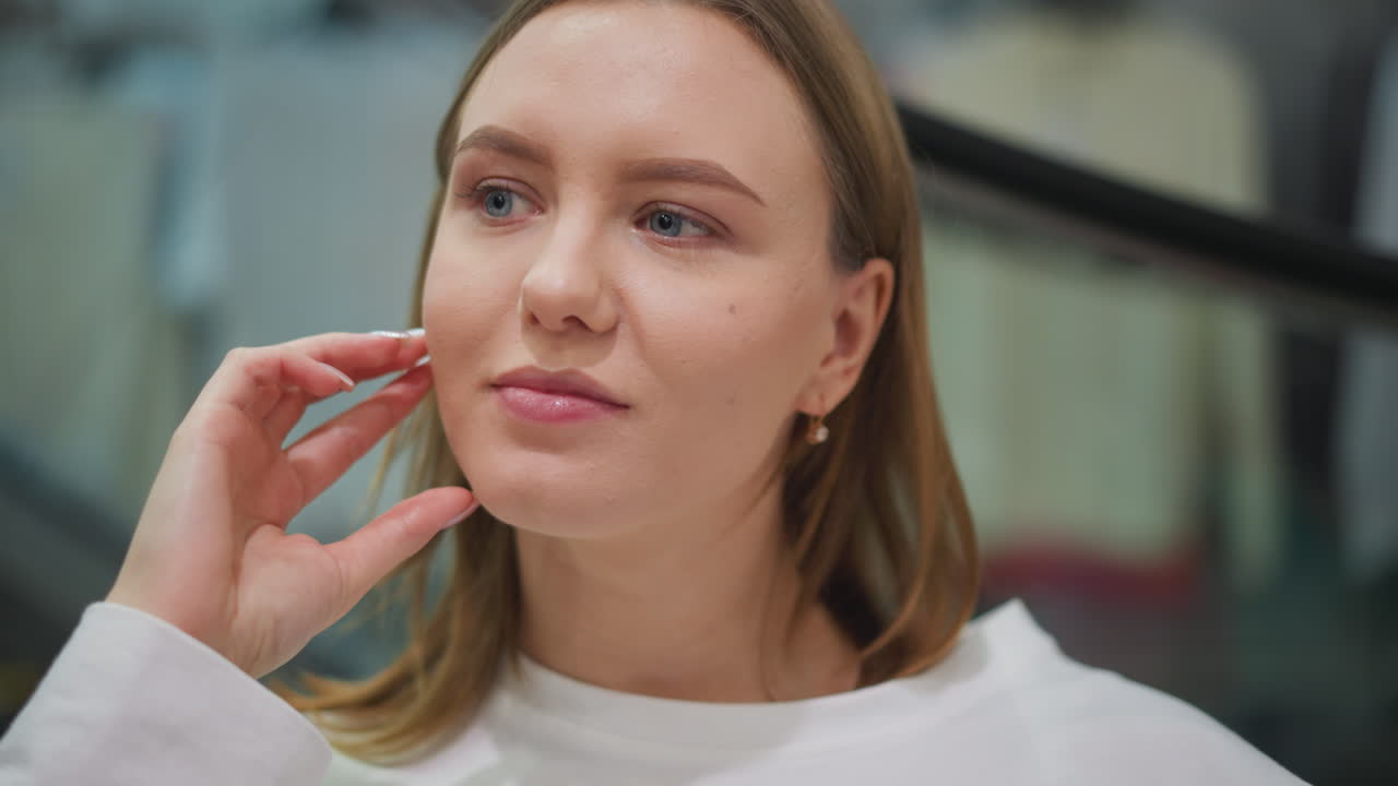 un primer plano de una mujer tocando pensativamente su frente en un centro comercial baja su mano con una iluminación suave, rodeada de exhibiciones de ropa en el fondo