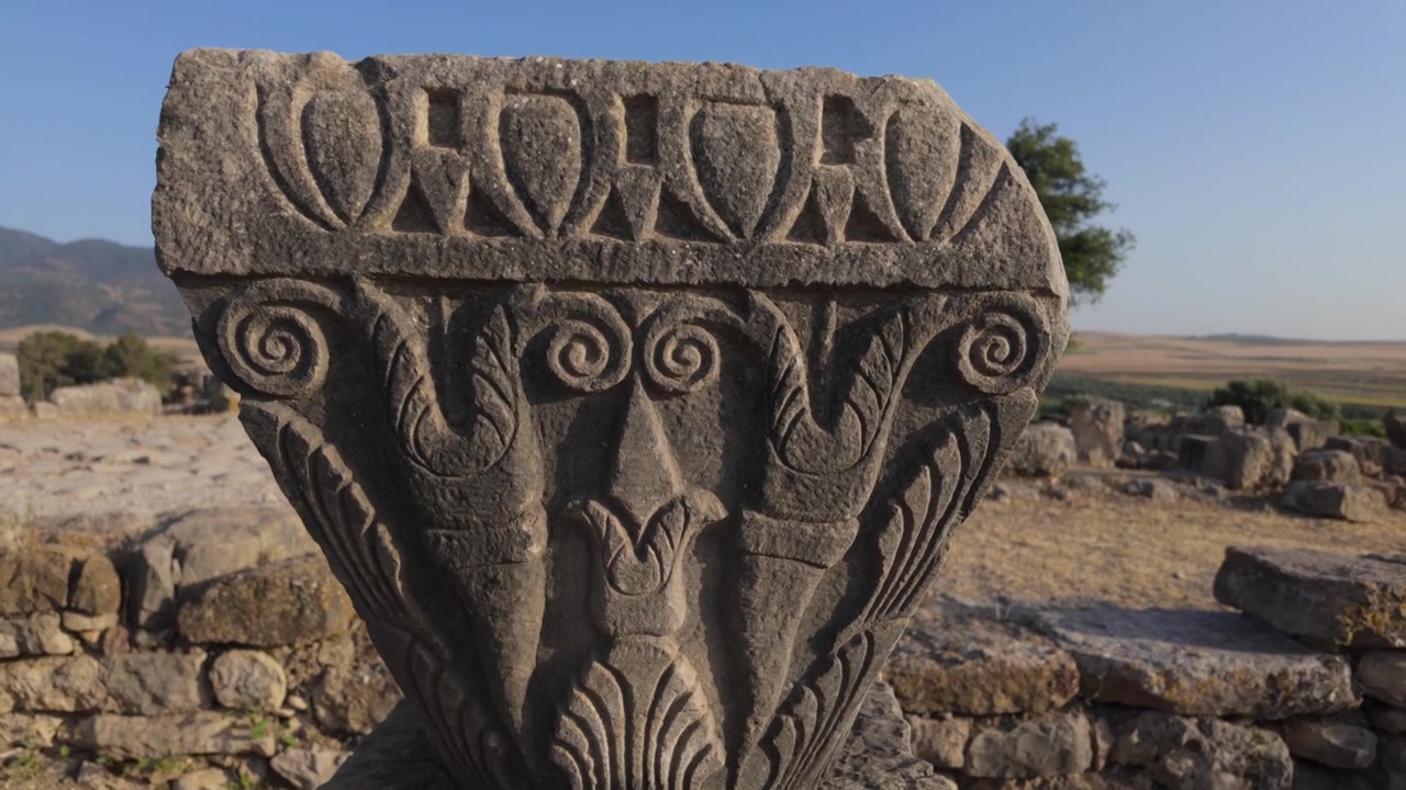 Fas'taki UNESCO listesindeki Volubilis arkeolojik alanında, klasik süsleme ve Latince bir yazıt içeren oyma bir Roma taşının yer seviyesinden sinematik görünümü.