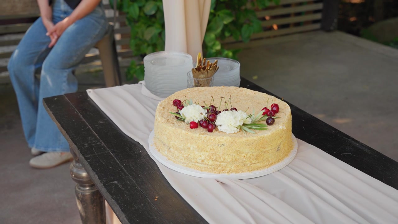 Una torta alla crema invitante, adornata con frutti di bosco freschi e fiori, è posizionata su un tavolo rustico nero all'aperto, con la luce del sole che filtra, mentre una persona vestita casualmente in jeans blu è seduta nelle vicinanze.