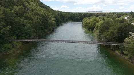 Aerial-drone-flight-over-river-with-tourist-crossing-Rainbow-Bridge-at-Kepler-Track-National-Park,-New-Zealand