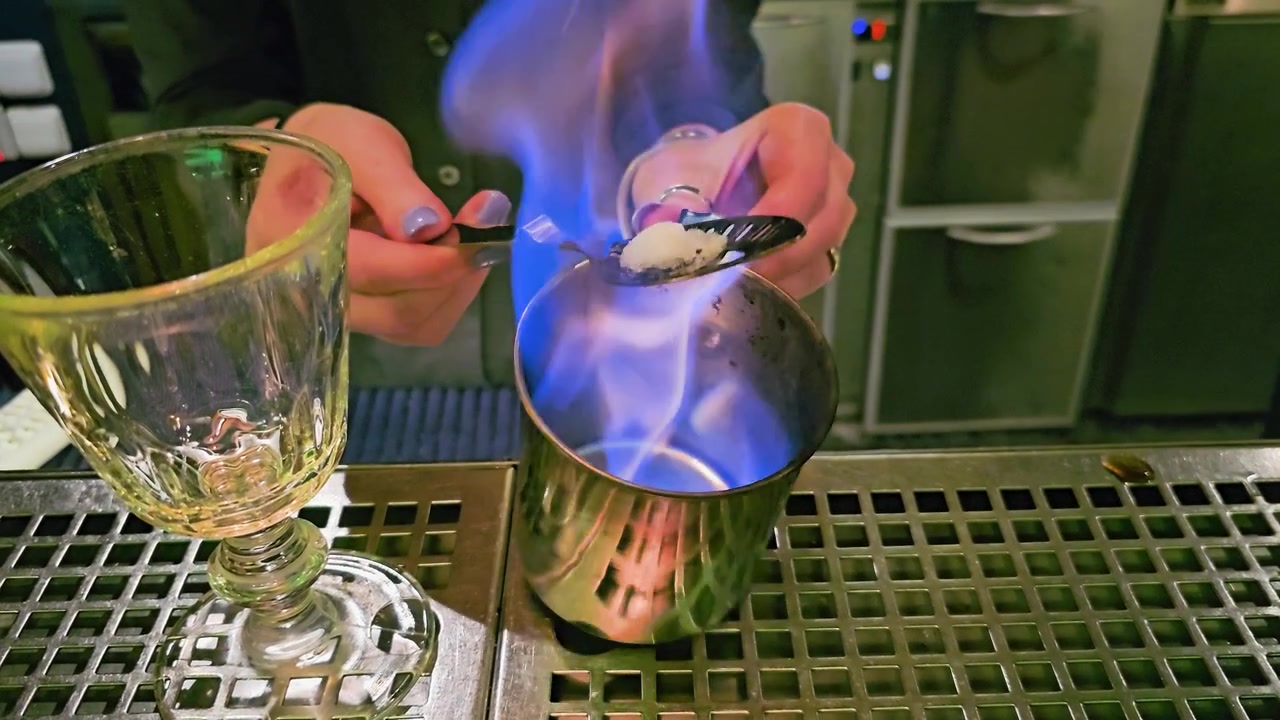 Primo piano di una preparazione di assenzio fiammeggiante su un bancone da bar in acciaio inossidabile, con zolletta di zucchero su cucchiaio forato, tazza di metallo, calice vuoto, mani del barista e illuminazione colorata da discoteca all'interno.