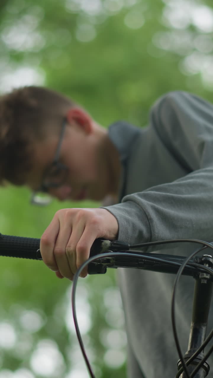 niño con gafas agarra firmemente el manillar de una bicicleta estacionada cerca de un árbol en un campo cubierto de hierba, está comprobando la funcionalidad de los frenos mientras está de pie, su cara está borrosa en el fondo