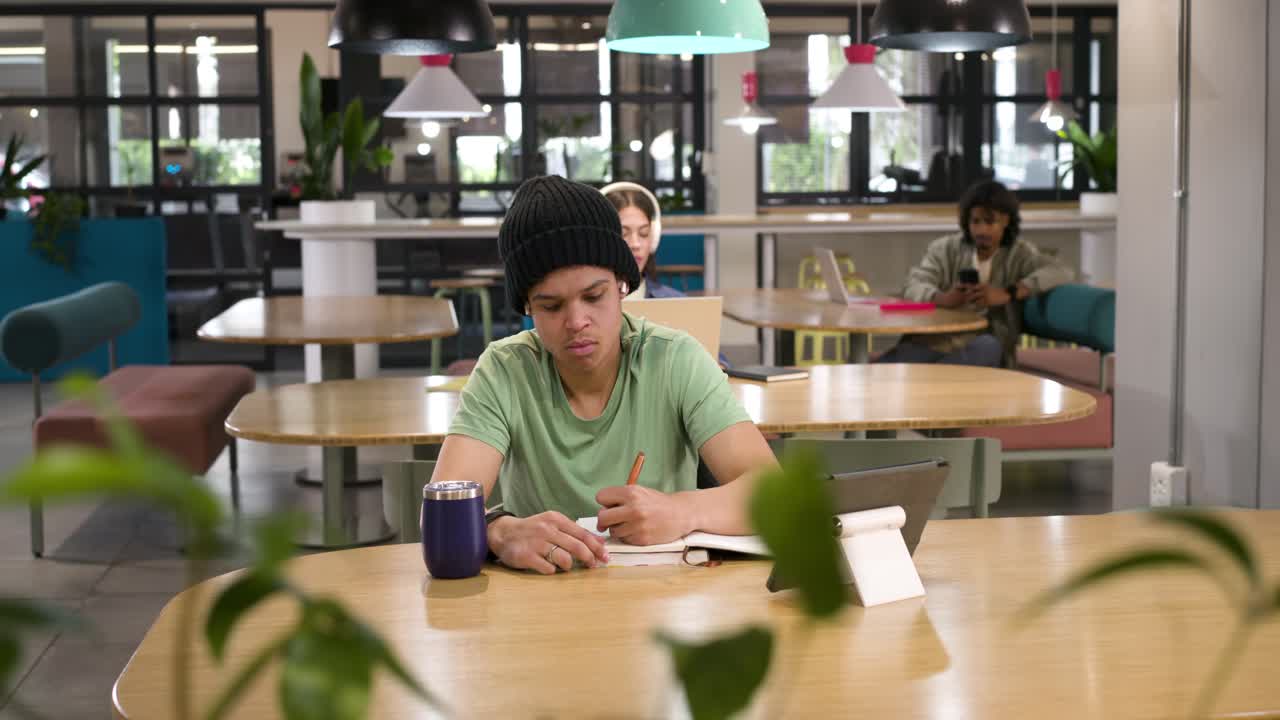 Studenti afroamericani che studiano, un ragazzo con il berretto che scrive su un quaderno e consulta il tablet.