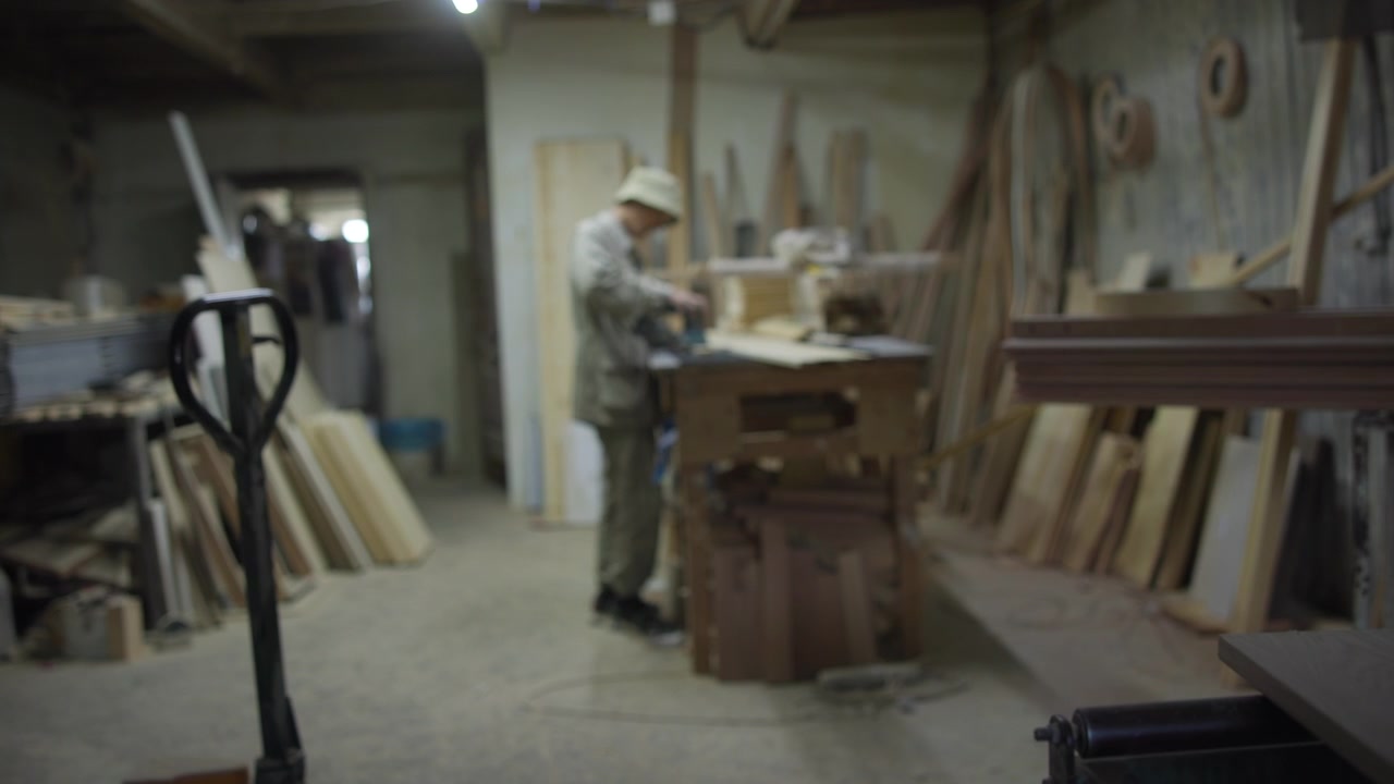 Carpenter sanding wooden panel at workshop workbench, pallet jack handle in foreground, cluttered lumber stacks and tools around under soft overhead light indoors