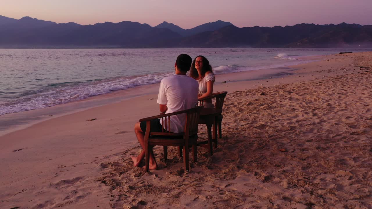 Young smiling couple sitting on chairs over sandy beach washed by sea waves at sunset with colorful sky over mountains horizon