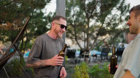 Over-his-shoulder-a-confident-guy-in-sunglasses-with-a-beard-talks-and-drinks-beer-from-a-brown-bottle-with-his-friend-in-the-courtyard-of-a-country-house