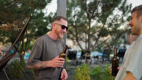 sobre su hombro un tipo confiado en gafas de sol con una barba habla y bebe cerveza de una botella marrón con su amigo en el patio de una casa de campo