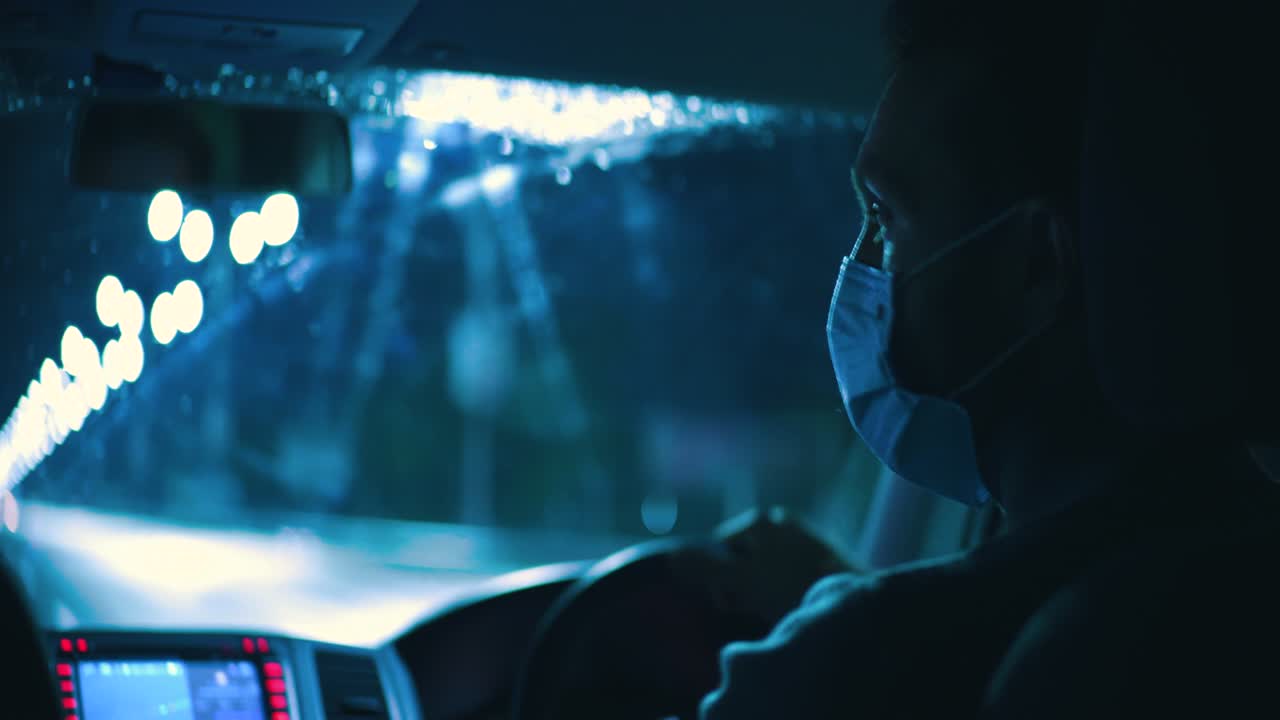 의료 마스크를 입은 젊은 남자가 밤의 비가 오는 고속도로를 통해 차를 운전합니다.