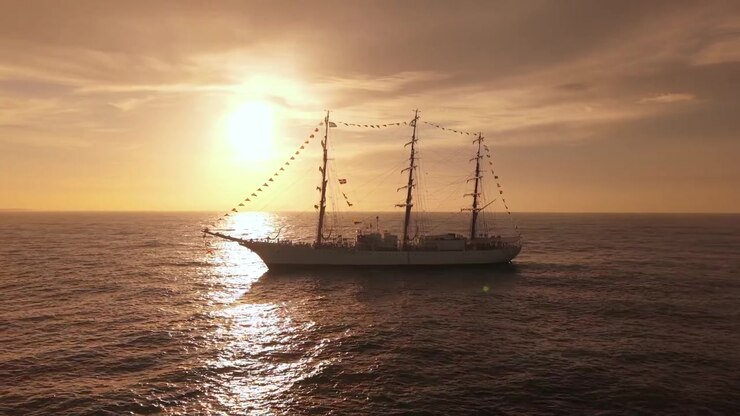 Aerial tracking shot of a majestic three-masted tall ship sailing on the open ocean at sunset, featuring heavy backlighting, lens flares, and golden sun reflections on the water