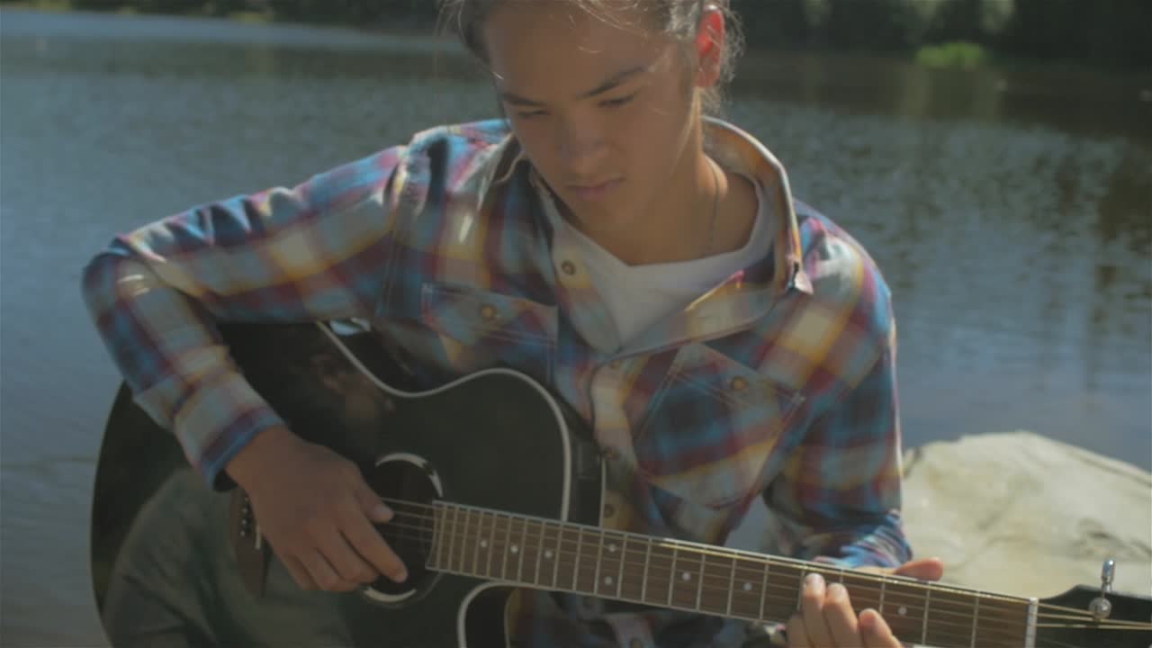Optagelse i medium slow motion af en ung asiatisk mand, der spiller musik på sin akustiske guitar ved en søbred
