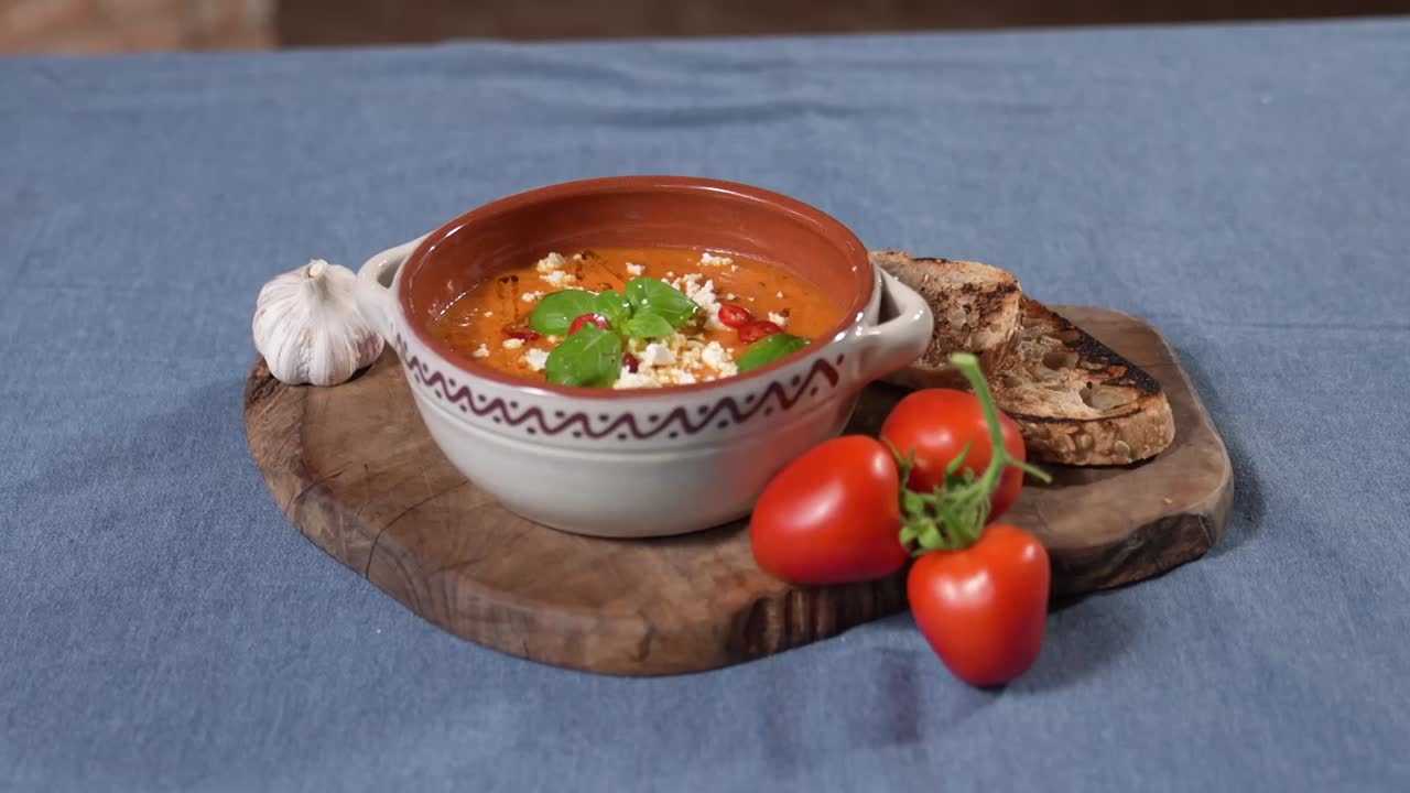 Traditional tomato soup served in a decorated ceramic bowl, garnished with herbs and cheese. Presented on a wooden board with fresh tomatoes, garlic, and toasted bread. A homestyle culinary setup