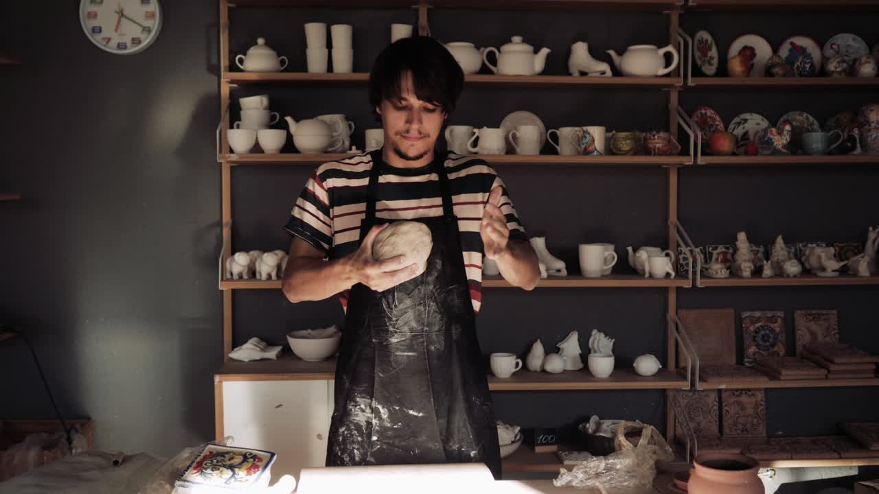 joven alfarero masculino amasa arcilla contra el fondo de un estante con espacios en blanco de cerámica