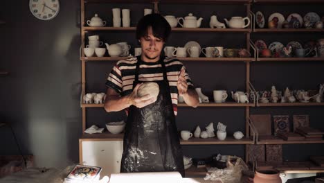 young male potter kneads clay against background of a rack with ceramic blanks