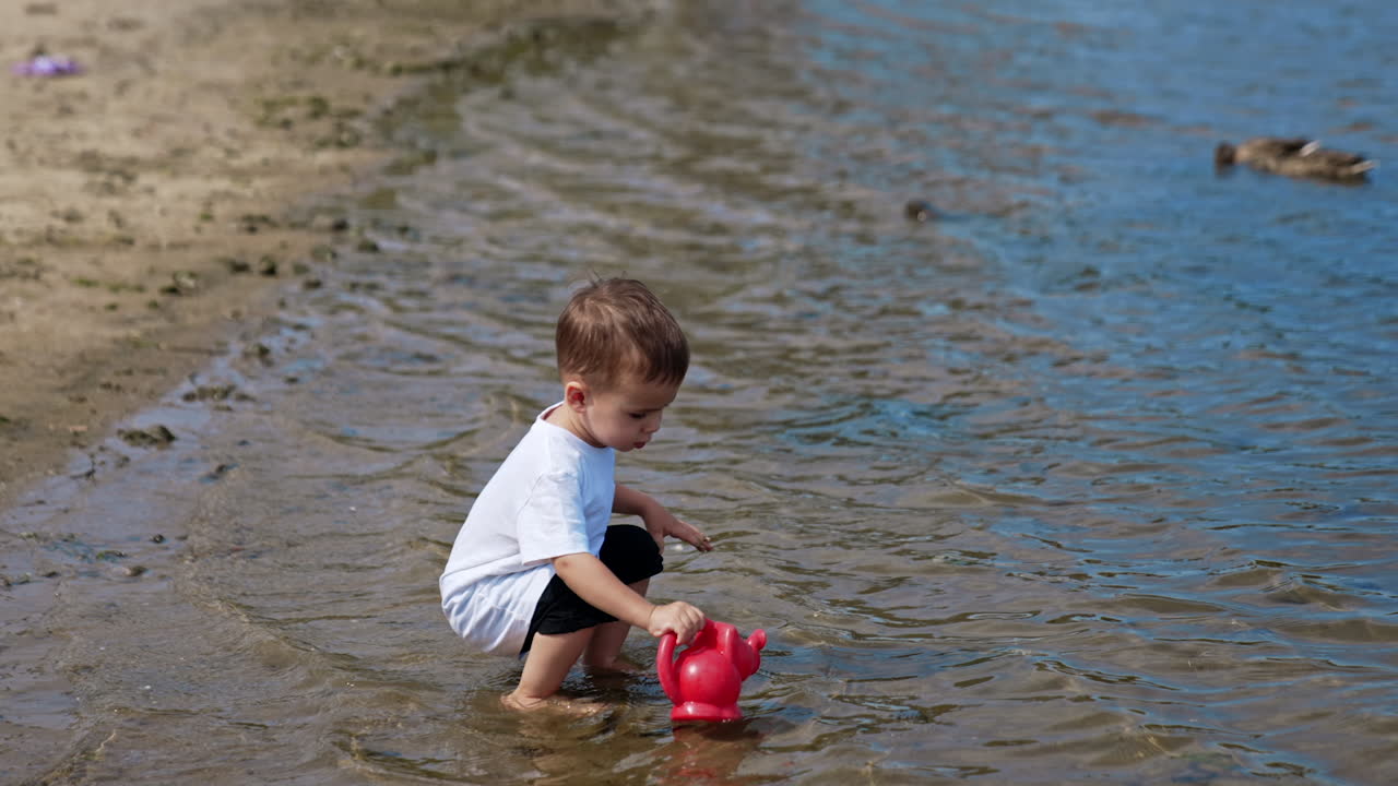 Lovely baby boy in white t-shirt walks to the seawater to fill his watering can. Caucasian child plays on the beach in summer.