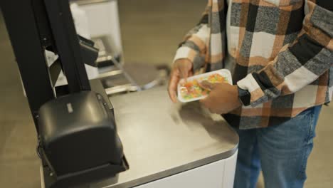 Close-up-shot-of-a-man-with-Black-skin-in-a-checkered-shirt-and-blue-jeans-scans-the-goods-he-needs-at-a-self-service-checkout-in-a-modern-large-grocery-store