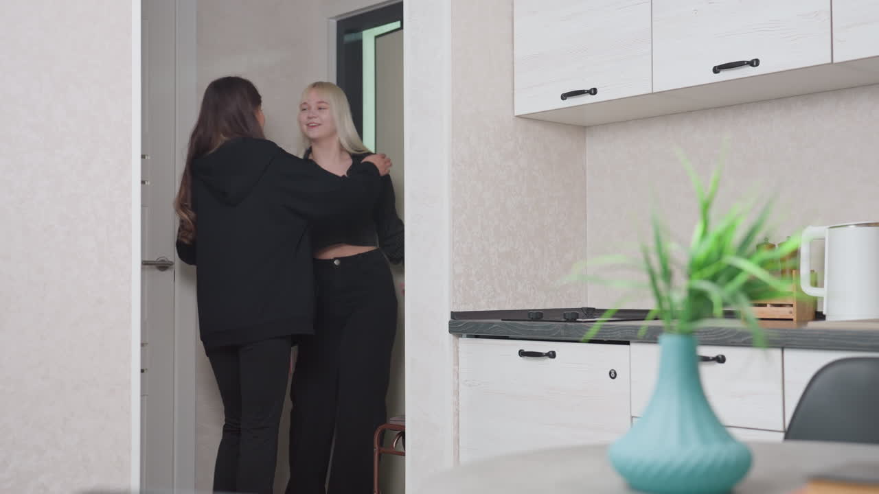 Young woman in black hoodie opens door and observes friend slipping on shoe in stylish hallway before heading out, capturing warm farewell moment between friends