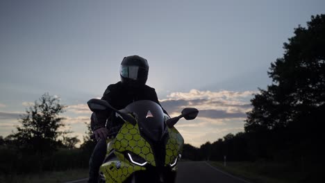 Motociclista-En-Moto-Bmw-Sport,-Joven-Motociclista-En-Moto-De-Moda-Se-Detuvo-En-La-Carretera-Asfaltada-En-El-Campo-Para-Descansar-Durante-El-Viaje-Al-Atardecer---Tiro-De-Cardán-En-órbita