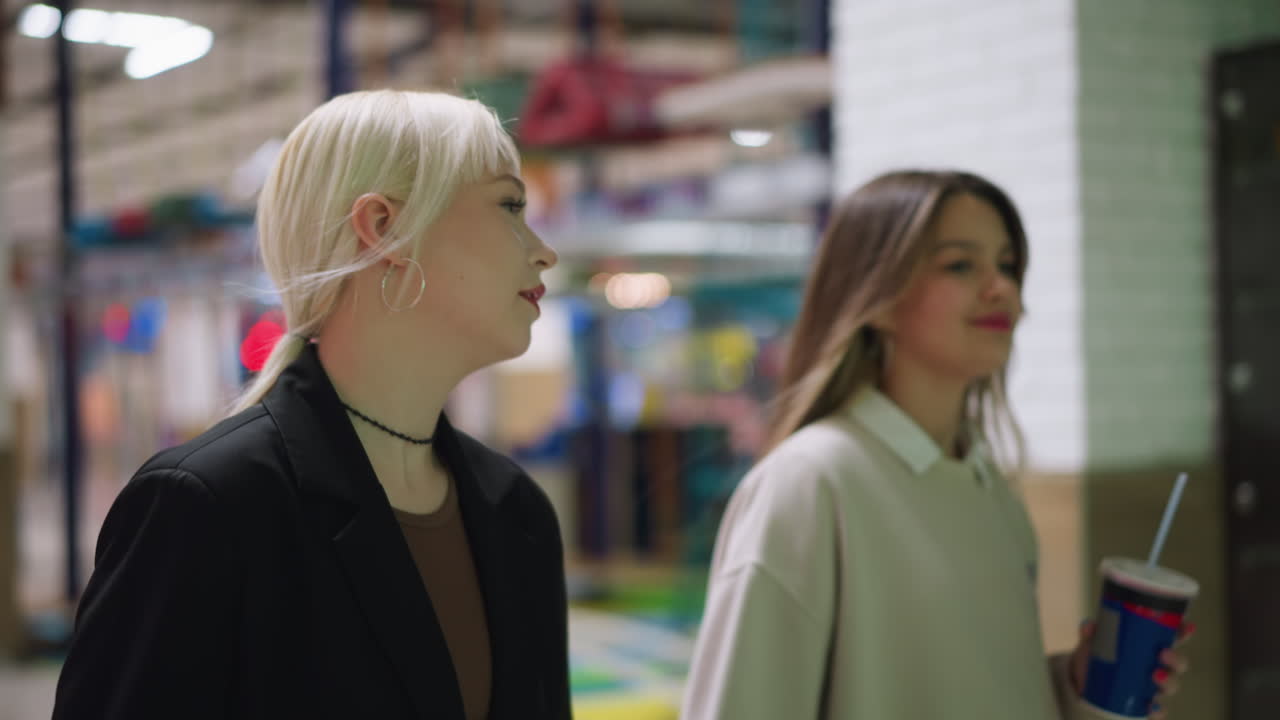 Colleagues walking together through mall corridor, one sipping cola drink while engaged in conversation, showing casual connection, stylish clothing and bright indoor environment