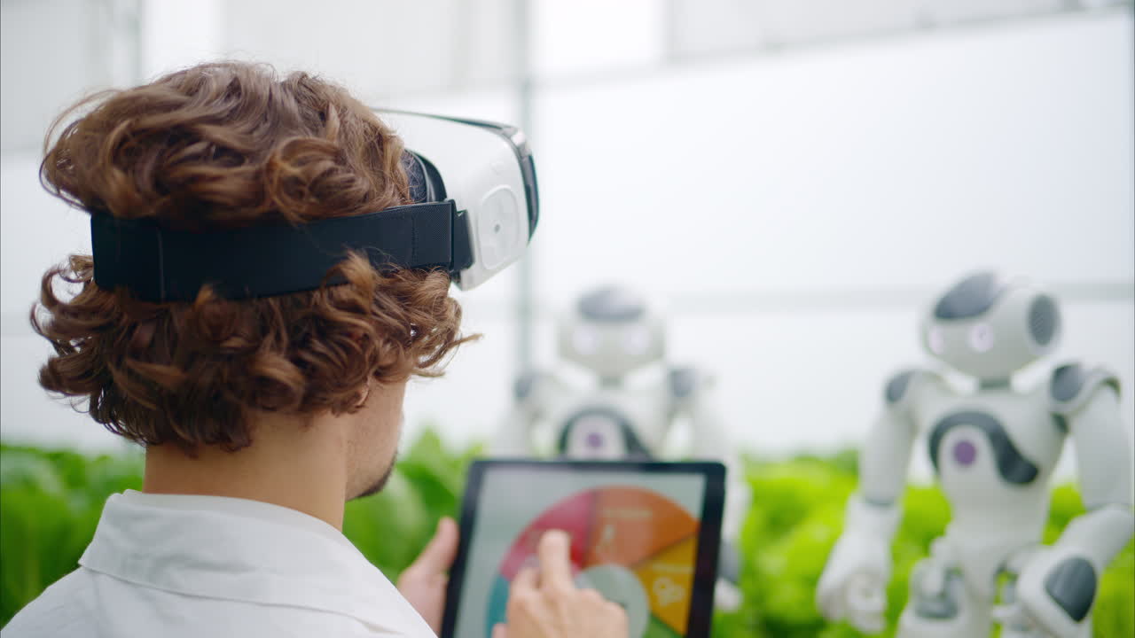 Laboratory technician in a white coat wearing virtual reality headset analysing graphs on a tablet while interacting with two humanoid robots in a greenhouse farm