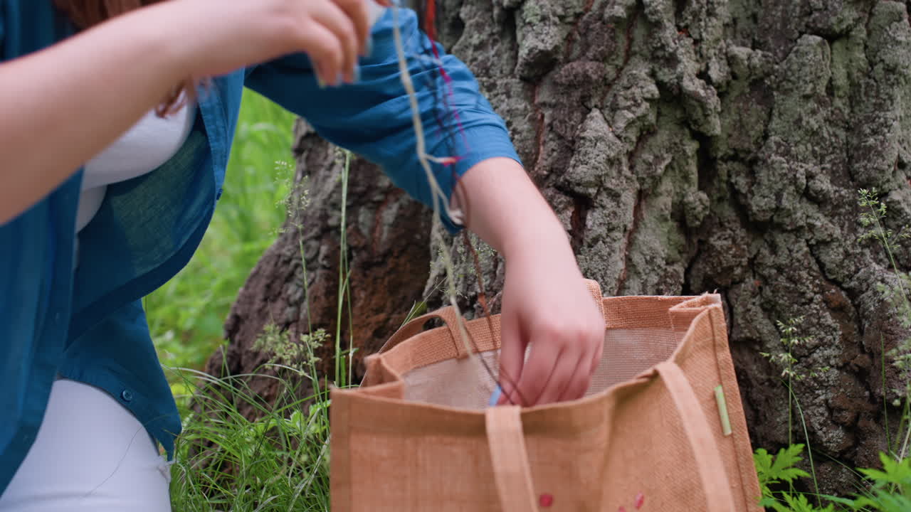 Close up of lady squatting beside large tree trunk reaching into brown fabric bag to get embroidery material during calm outdoor crafting moment surrounded by green grass