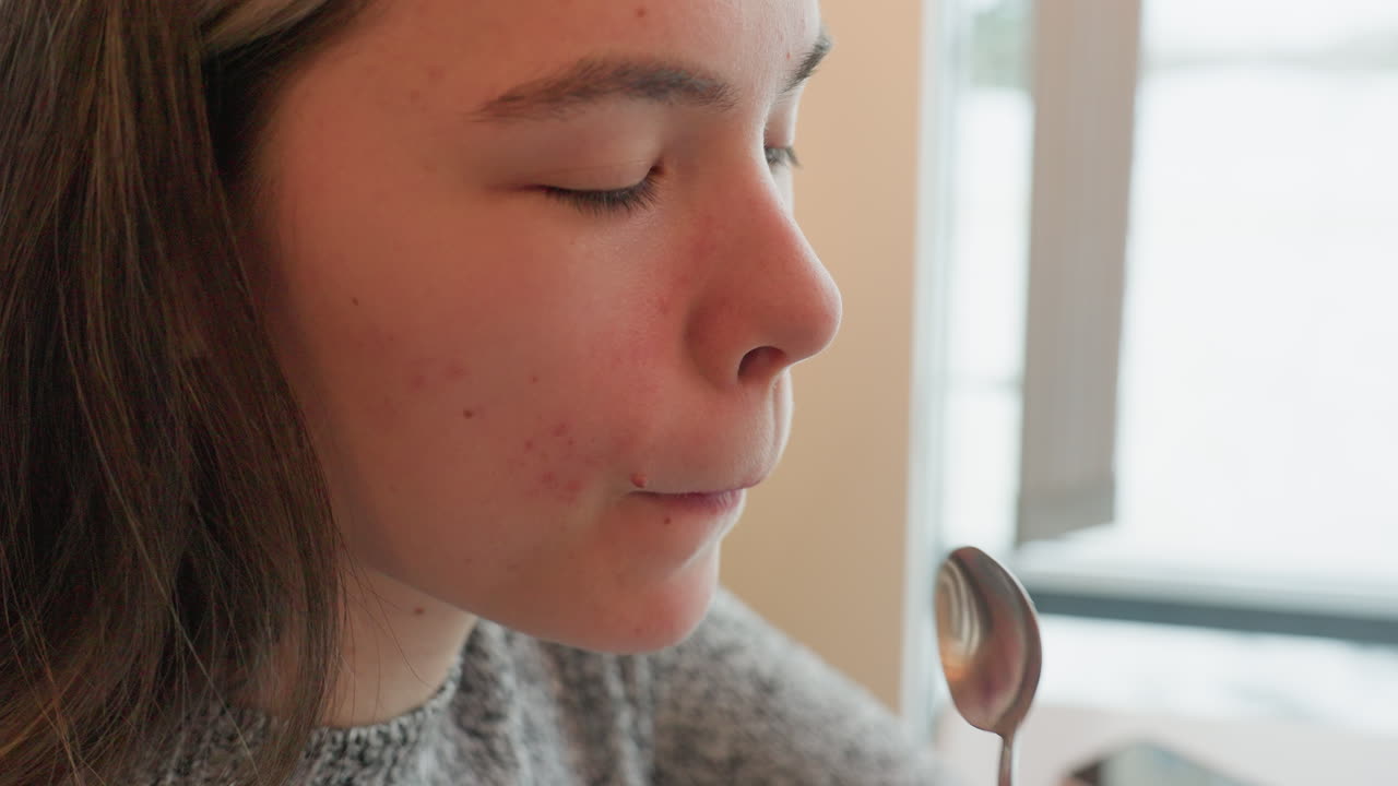 Fair tall girl seated in cafe scooping tea with teaspoon enjoying satisfying taste expression on face shakes head in approval cozy setting morning light streaming through window adds warmth