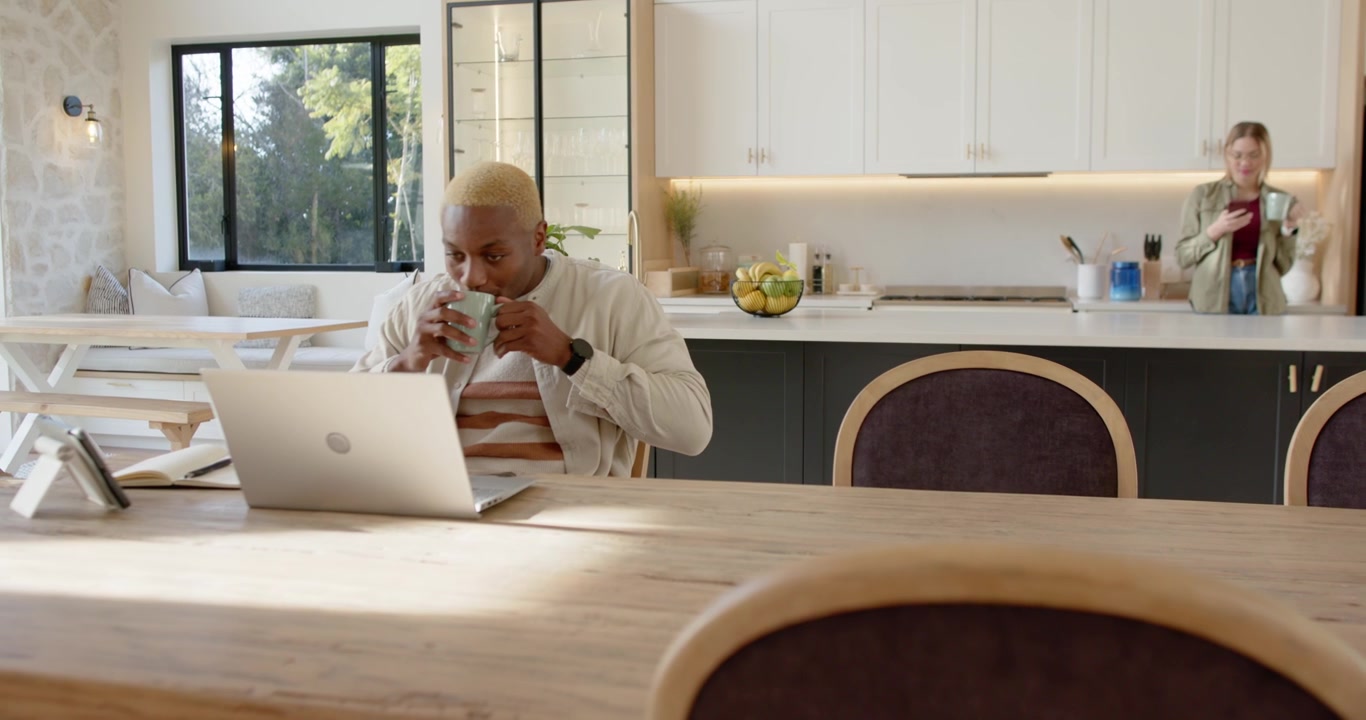 African American couple sharing kitchen, camera zooming on man sipping mug and working on laptop