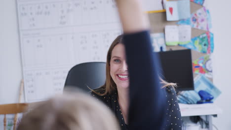 Young-female-teacher-sitting-at-desk,-children-raising-their-hands-during-a-lesson,-selective-focus