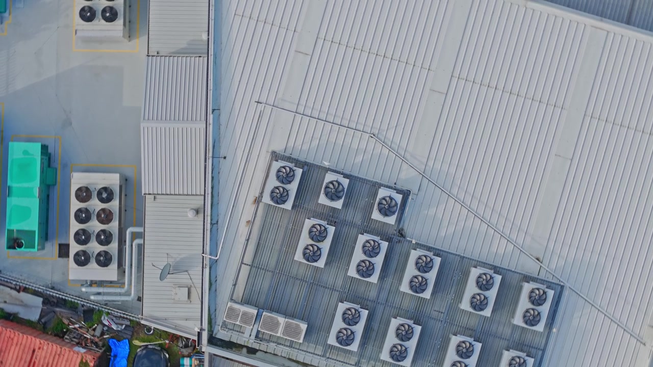 High-altitude drone footage captures a modern data center roof lined with industrial HVAC cooling units, vents, piping, and steel structures in bright, even daylight. No people visible.