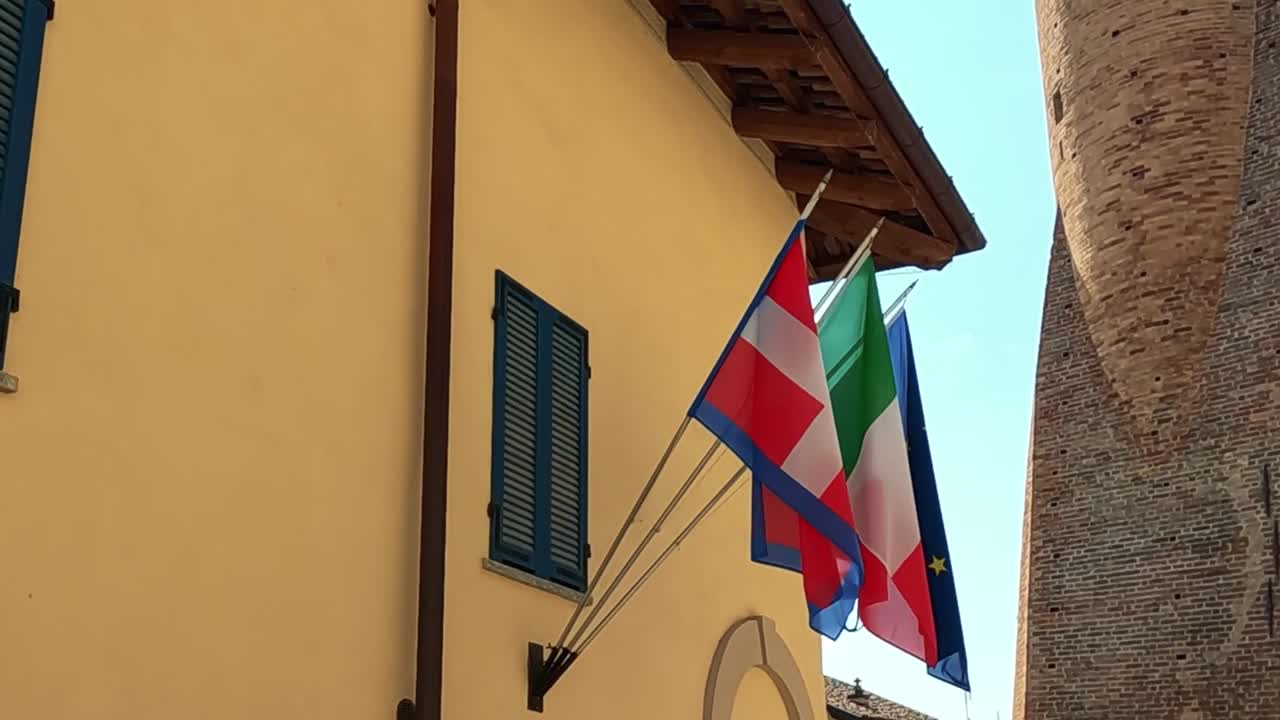 Close-up of Italian and EU flags on a sunlit building facade with blue shutters.