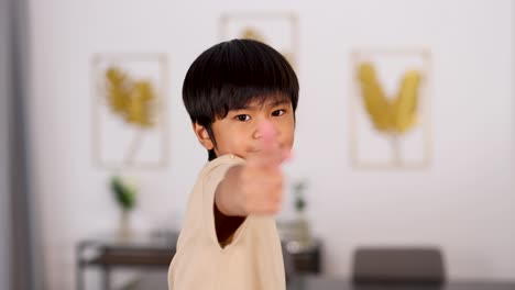 young asian boy in beige shirt smiles, points, and gives a thumbs up in a bright, modern room with soft lighting and minimal decor