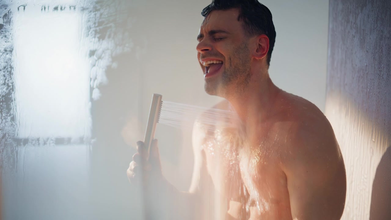 Cheerful man singing in shower holding showerhead mic. Close up wet male in bath