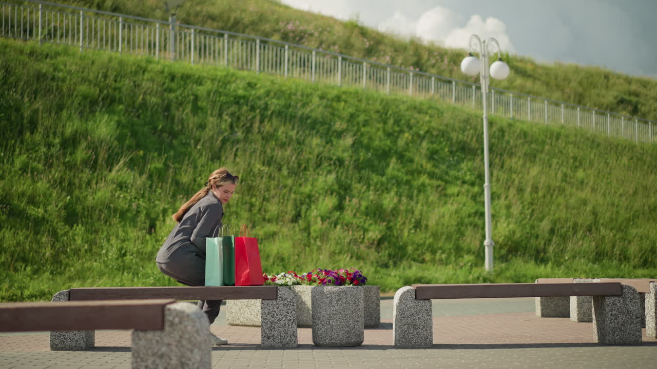 한 여인이 벤치에 다가와, 그 위에 다채로운 쇼핑 가방을 떨어뜨리고, 앉아서, 왼손을 벤치 위에 놓고 멀리 바라보며, 배경에는 언덕의 초록색이 있습니다.