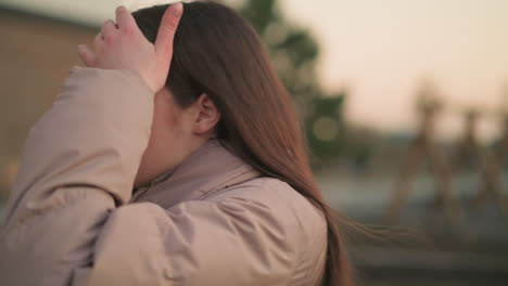 this close-up shot captures a young woman in a peach jacket, walking through a park with a thoughtful expression. her hand rests on her forehead, suggesting contemplation or perhaps fatigue