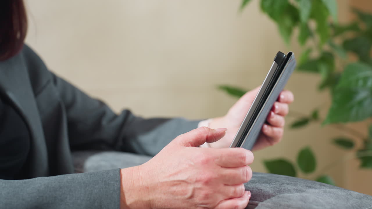 Businesswoman in suit sitting comfortably while using tablet indoors near green plant, focusing on screen for digital task, online communication, or remote work in relaxed professional environment