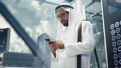 successful muslim businessman in traditional white kandura riding glass elevator to office in modern business center. man using smartphone. saudi, emirati, arab businessman concept.