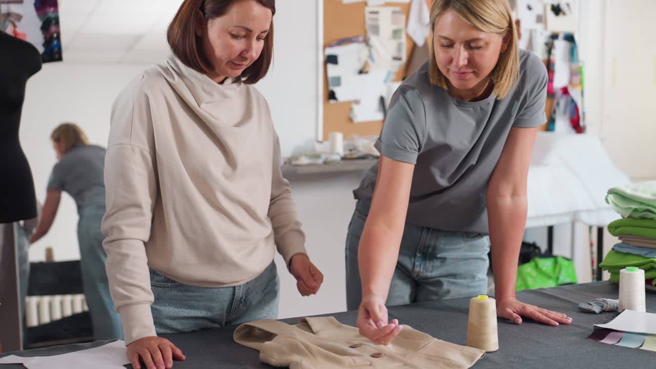 Two stylists bend over beige blazer on worktable, debating ideal button placement, testing positions by hand while mirror, mannequin, pattern scraps, thread spools surround fashion workspace