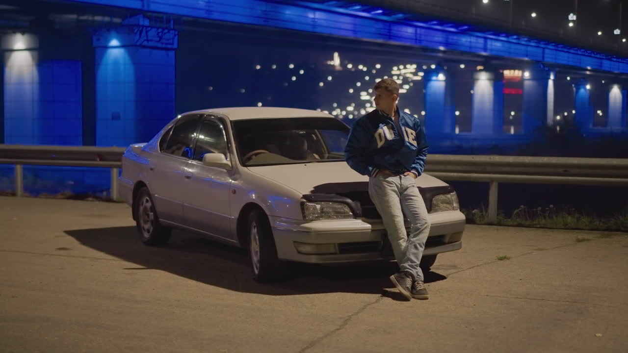 puente nocturno, aparcamiento, hombre apoyado en un sedán clásico bajo luces azules, manos en los bolsillos, reflejos en el capó, asfalto tranquilo, bokeh distante de la ciudad, pausa introspectiva, luz baja cinematográfica, grano sutil