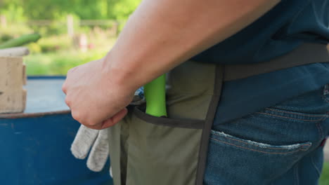 rear view farmer in denim and garden apron stowing shovel and hand glove into pocket beside blue drum, garden background, daylight closeup, rural work prep, practical gear