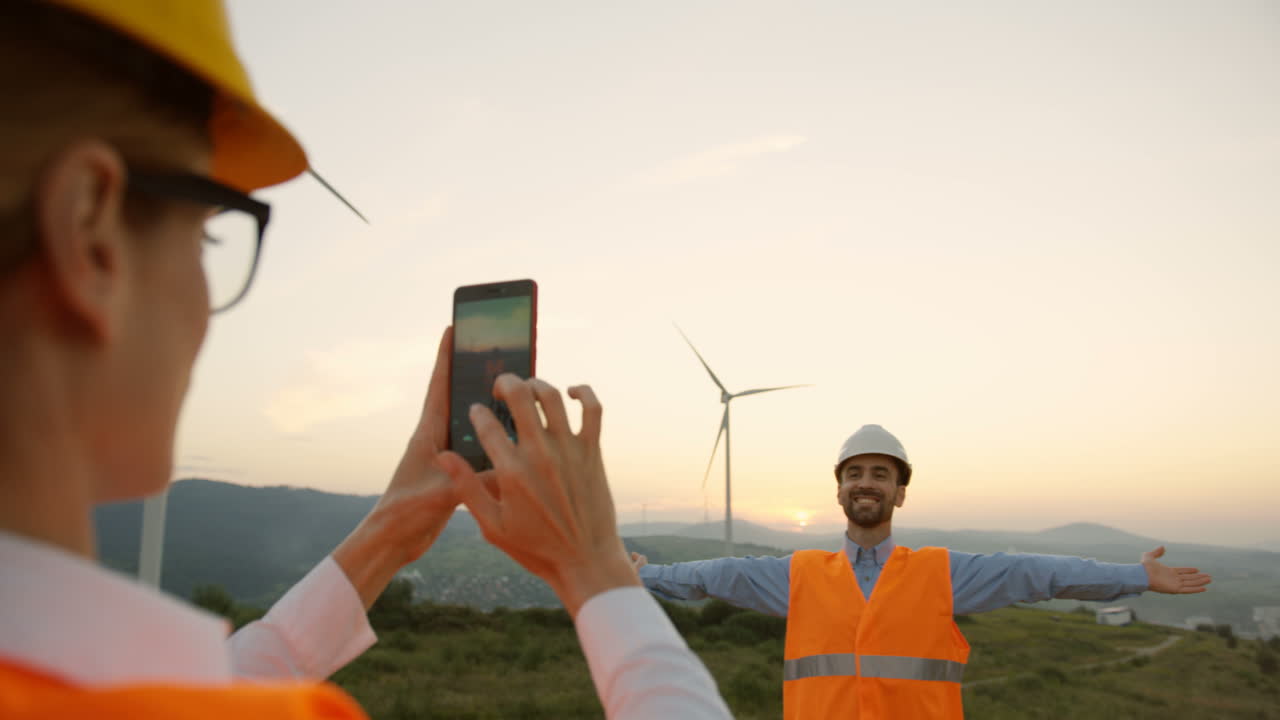 ingeniera caucásica con casco tomando una foto a su colega con un smartphone en las enormes turbinas de molinos de viento al atardecer