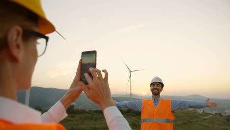 caucasian female engineer wearing a helmet taking a photo to her colleage with smartphone at the huge windmills turbines at sunset