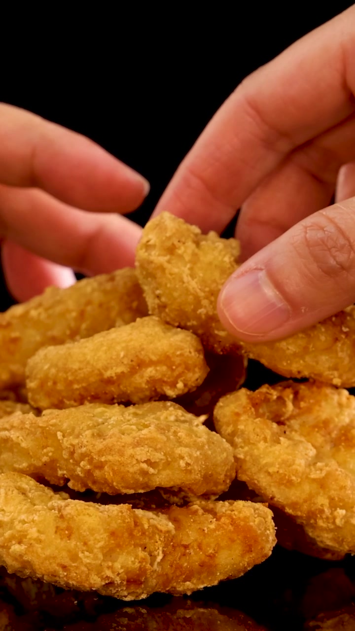 Primer plano vertical de manos tomando una pila de nuggets de pollo dorados sobre fondo negro.