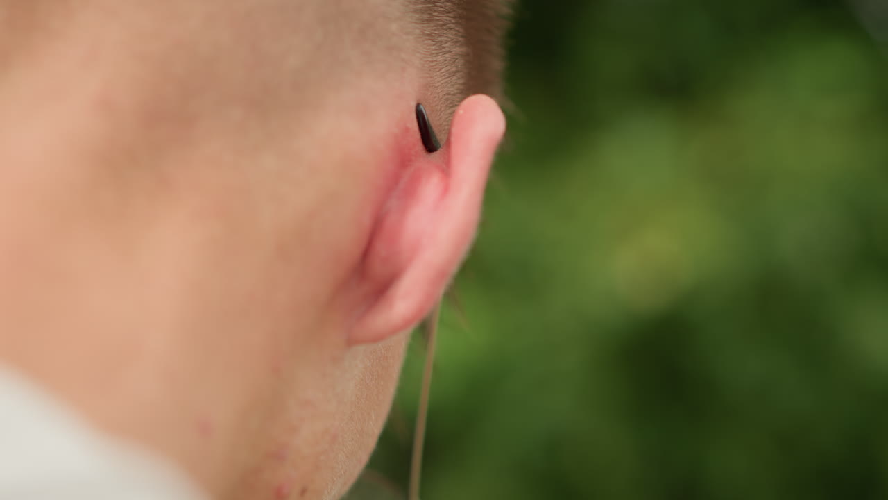 Studente che si toglie l'auricolare, giovane caucasico che si sistema l'auricolare con un dito in un ambiente esterno, studente con la nuca rasata che indossa una felpa con cappuccio mentre manipola l'auricolare su uno sfondo bokeh verde e suggestivo