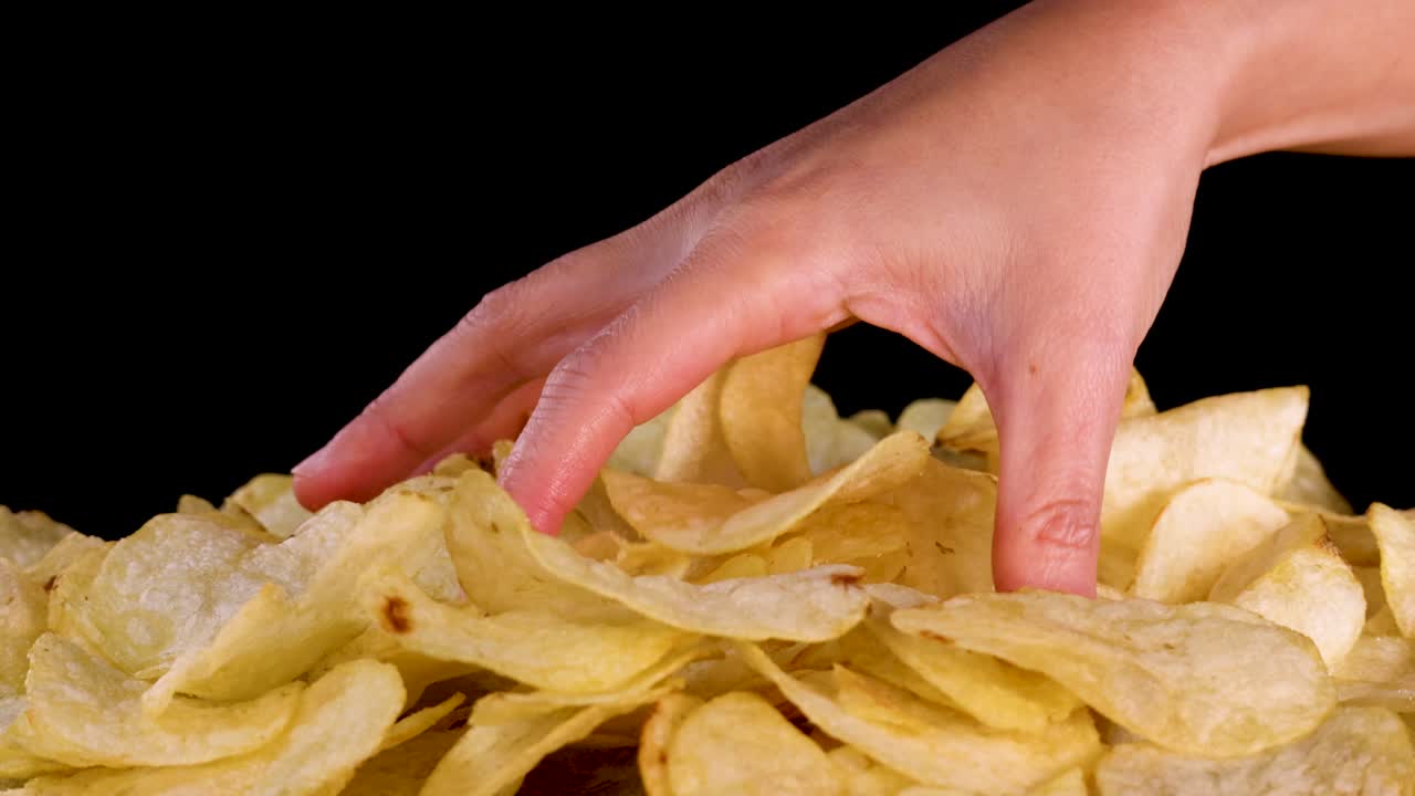 Human hand picks potato chips from large pile, close-up, studio lighting, black background