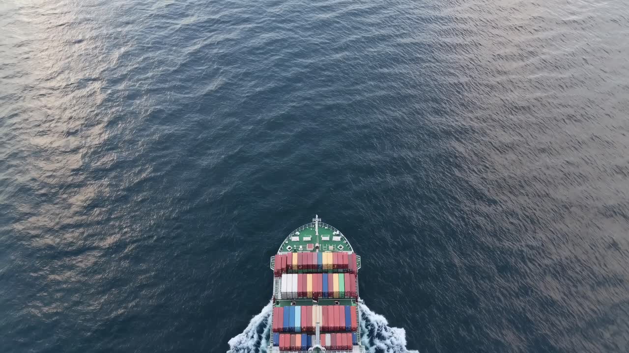 Container ship traversing calm blue ocean waters, creating rippling wake, carrying colorful cargo containers visible from aerial perspective, representing global maritime shipping logistics