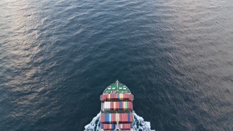 container ship traversing calm blue ocean waters, creating rippling wake, carrying colorful cargo containers visible from aerial perspective, representing global maritime shipping logistics