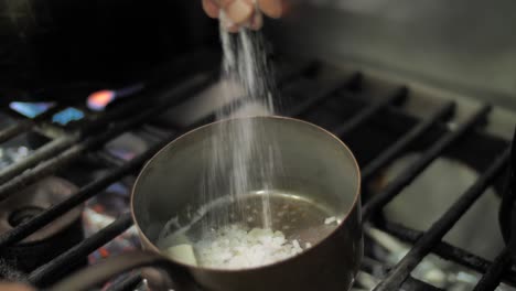 chef adding salt to frying onion and butter in metal pot on gas stove