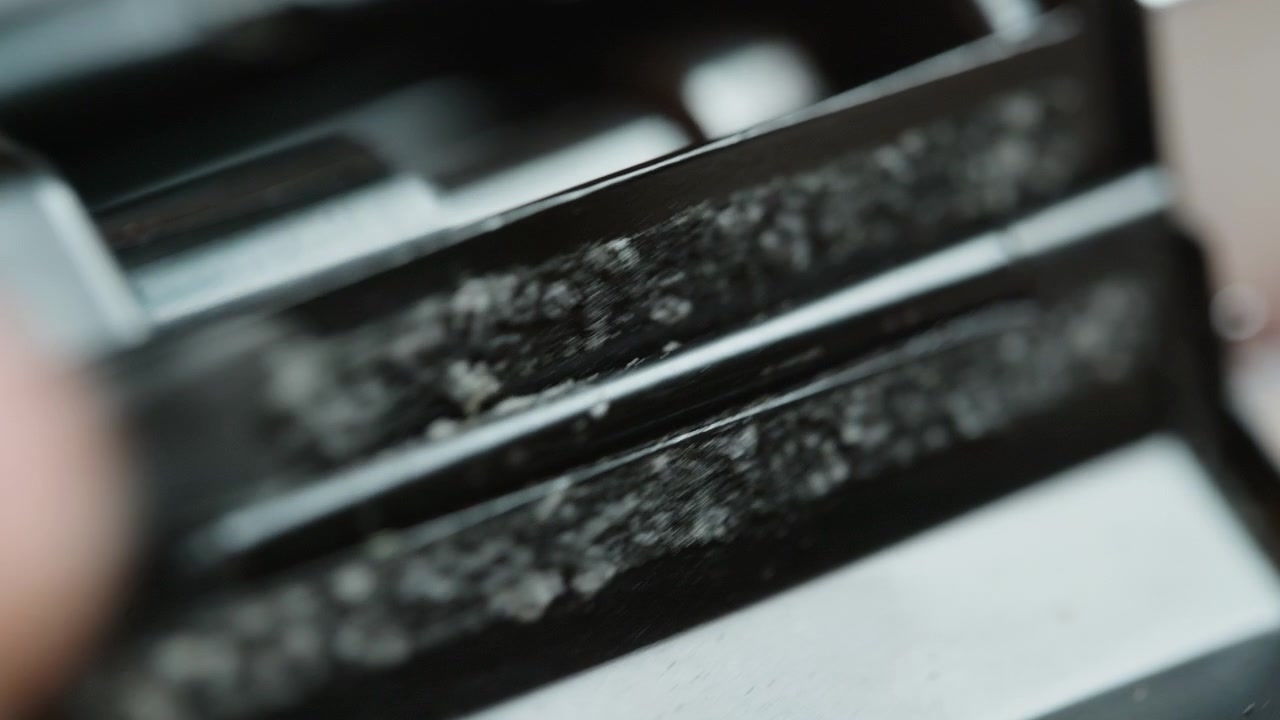 Macro shot: the vintage 35mm camera is repositioned, then a tool scrapes crumbled old light seals from the film channel, foam debris breaking away. Detailed restoration of analog gear before renewal.