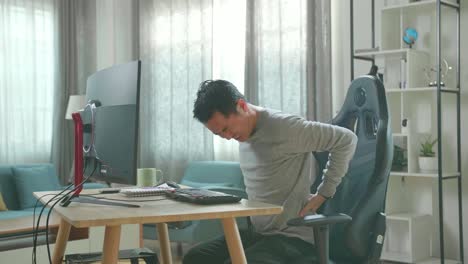 hombre asiático con camiseta de mangas largas y pantalones negros con dolor de espalda mientras usa una computadora de escritorio para trabajar en casa.