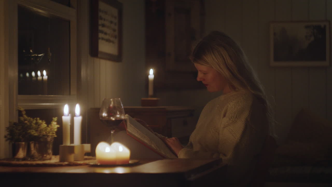 A woman in a knit sweater writing in her journal by candlelight in a cozy cabin, surrounded by warm decor and a glass of wine.