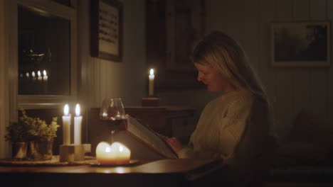 a woman in a knit sweater writing in her journal by candlelight in a cozy cabin, surrounded by warm decor and a glass of wine.
